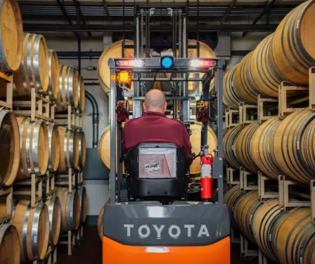 Forklift operator driving a Toyota electric forklift near pallet racking with barrels.