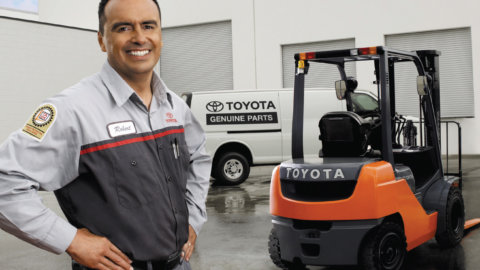 A male forklift service technician poses outside of the warehouse near an orange Toyota forklift.